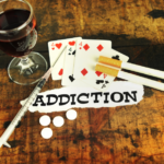 Photo qui symbolise plusieurs types d'addictions : 2 cigarettes, un verre de vin, un jeu de cartes, une seringue, le mot "addiction ..." inscrit sur un papier.