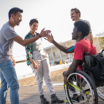 Photo d'un groupe de 4 jeunes adultes composé d'un homme en fauteuil roulant. Il tape la main d'un des adultes. Tous les adultes rient.